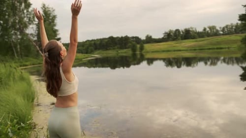 Young Woman Stretches Her Arms Up Doing Yoga Against the Background of a Pond