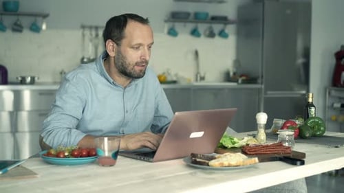 Man Works on Laptop at Kitchen Table with Food