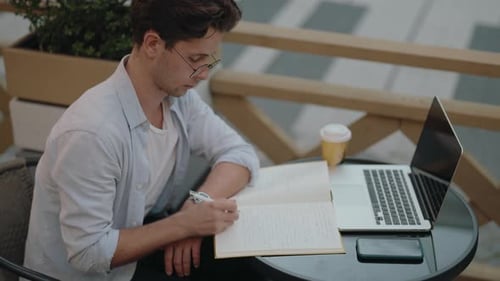Young Man Working Remotely on Laptop and Writing in Notebook at Outdoor Cafe