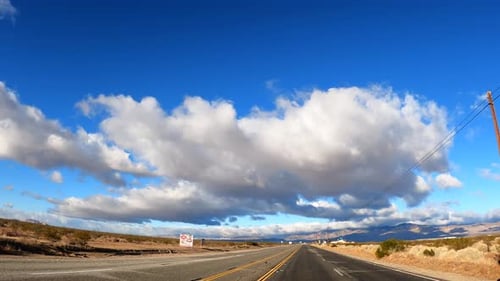 Driving through the Mojave Desert with large cloud formations overhead and the rugged mountains in t