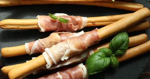 Grissini breadsticks with prosciutto and basil on wooden table, spinning. Top view