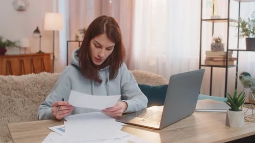 Focused Woman Freelancer Reading Documents and Using Laptop Computer at Table in Living Room at Home