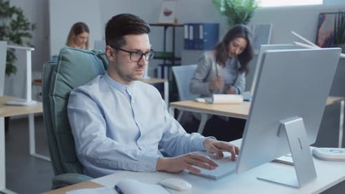 Attractive Young Man with Glasses Working on Computers in the Office In the Background
