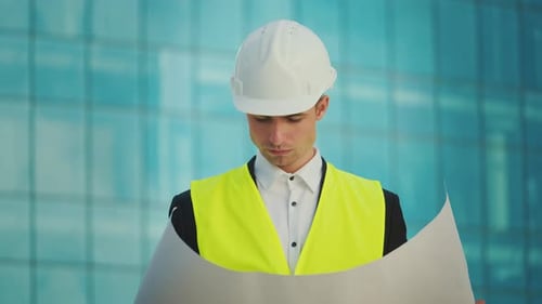 Male Engineer Wearing in Helmet and Safety Jacket Looking Construction Drawings in His Hand