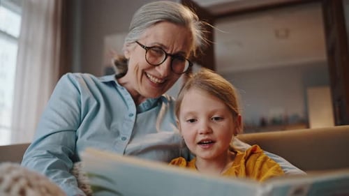 Close Up of Little Girl Reading Book Together with Her Elderly Loving Grandmother in Living Room