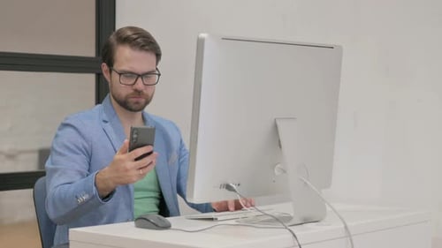Man Working at Computer While Using Smartphone