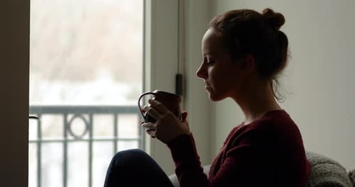 Medium shot of young woman quietly watching snowflakes fall outside and enjoying hot beverage in mug