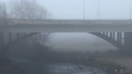 cars passing over a mysterious bridge with a lot of fog and a river below it