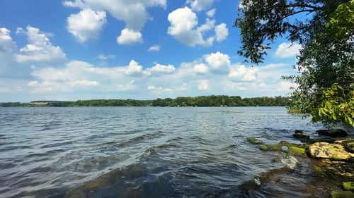 Little waves roll to the rocks on the river bank. Beautiful waterscape under blue sky with clouds