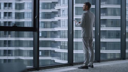 Confident entrepreneur stands thinking in modern office browsing tablet device