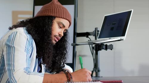 Man picking up pen and writing notebook notes after reviewing laptop charts at desk, copy space
