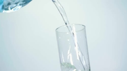 NAHAUFNAHME IN ZEITLUPE Füllen Sie ein transparentes Glas mit sauberem Wasser aus einer blauen Flasche auf weißem Hintergrund