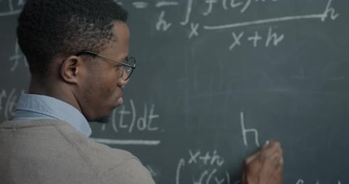 African American Physics Teacher Writing Equation on Chalkboard Teaching in Class