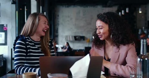 Two Beautiful Happy Smiling Young Multiethnic Friends Women Talk Laugh and Use Laptop at Modern
