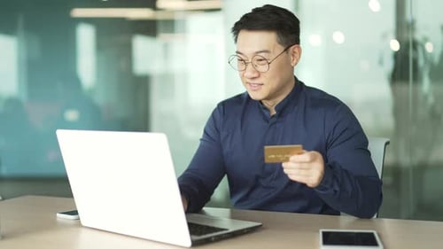 Smiling asian IT programmer employee paying with credit card on laptop at the office workplace Happy
