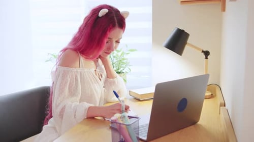 Young Woman Writes at Desk Indoors With Laptop