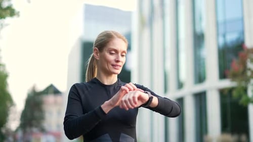 Happy smiling woman runner looking smart watch standing in city street. Sportswoman using checking