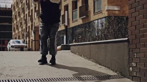 A Young Man Dressed in Casual Streetwear Walks and Bends in an Urban Alleyway with a Highrise
