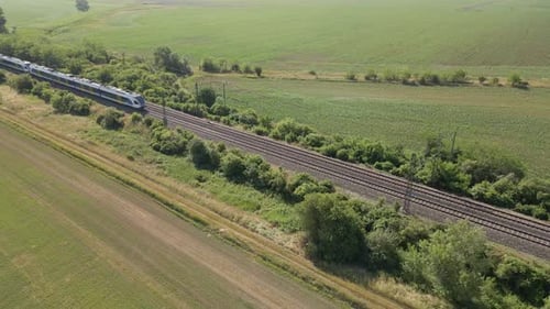 A modern passenger train moves along dual railway tracks through a lush green rural landscape. The s