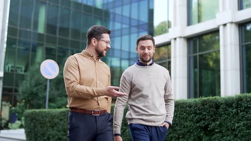 Two businessmen walk outside near an office building, having a cheerful, friendly conversation,