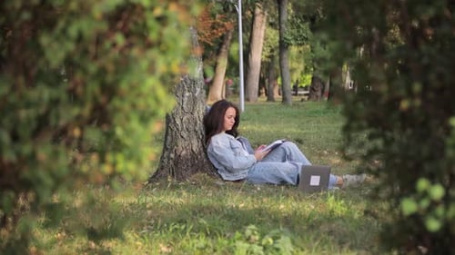 Eine junge Studentin sitzt mit einem Computer auf der Wiese im Park und studiert an der Universität