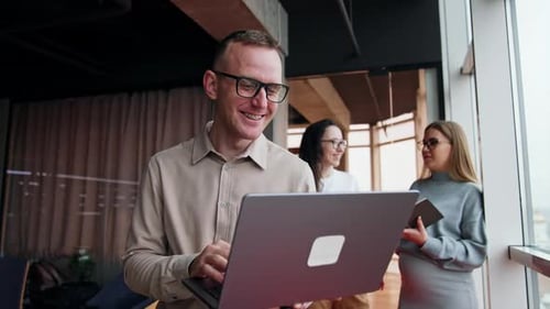 Lachender kaukasischer Mann mit einem Laptop. Kolleginnen diskutieren im Hintergrund über Arbeitsfragen.