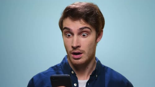Excited young man surprised browsing internet news on smartphone in studio slow motion