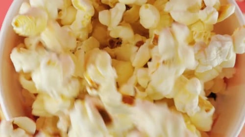 Granos de palomitas de maíz que caen en un balde de rayas rojas y blancas aislados sobre fondo rosa.