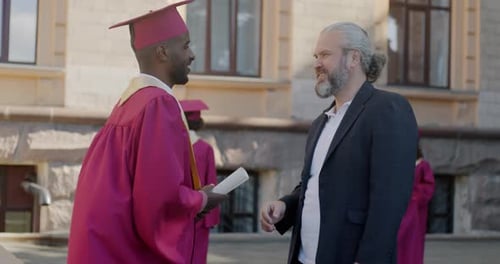 College Teacher Talking to Graduate Student in Mortar Board and Gown Shaking Hands and Hugging Man
