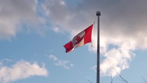 Canadian flag fluttering in the wind on a tall flagpole with white clouds in the background on a bri