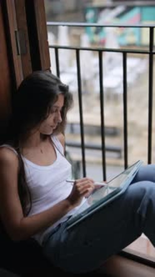 A Young Woman is Drawing on a Tablet By a Window Exuding Casual and Relaxed Vibes