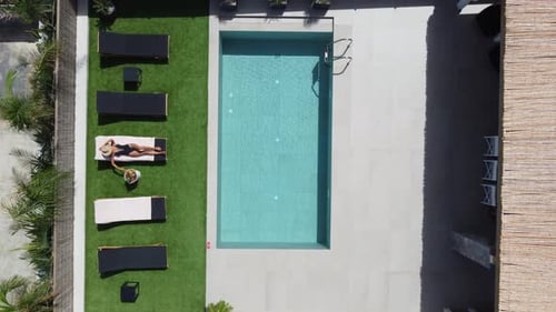 Drone flying over an outdoor pool in bird's eye view - Woman in swimming costume lying on sunbed and