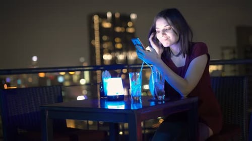 Young woman browsing internet on smartphone at night in rooftop bar