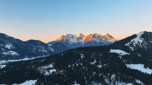 The drone is flying over a mountain with trees towards the big mountain lit by the golden sun during