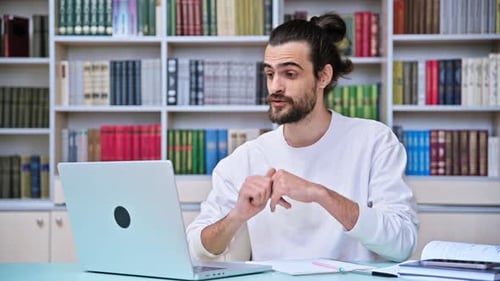 Young Man Talking on Video Call Using Laptop Sitting in Office with Book Shelves