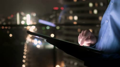A Man in a Suit at Work Reads the News on a Tablet Outside the Window the Glowing Windows of