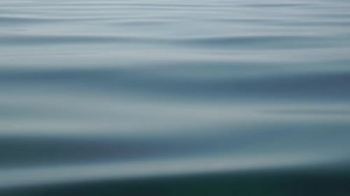 Sea Water Surface Camera Flies Over the Calm Azure Sea with Volcanic Rocky Shores on Background