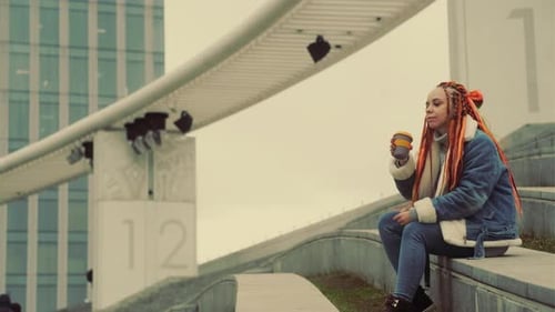 Young Woman with Dreadlocks Drinking Coffee From Cup Looking Away Sitting on Concrete in Modern City
