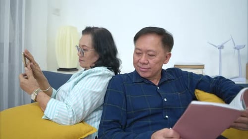 On a holiday, an elderly couple relaxes at home reading a book and chat on mobile phone