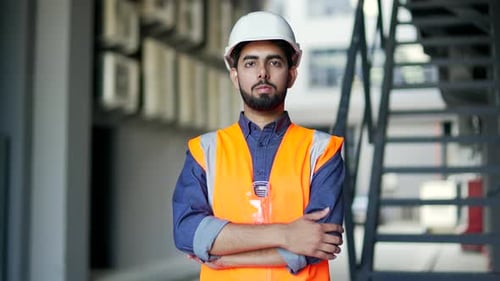 Portrait of confident young professional engineer wearing safety helmet and vest standing at factory