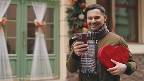 Handsome Caucasian Man Standing in the Winter Decorating Street Holding Heart Shape Gift and Using