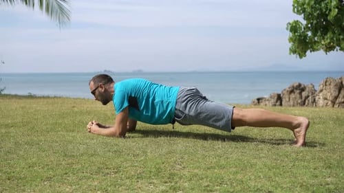 Junger Mann macht Plankenübungen zur Stärkung der Rumpfmuskulatur in einem sonnigen Garten am Meer