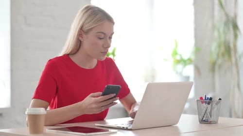 Young Blonde Woman Browsing Internet on Smartphone while using Laptop in Office