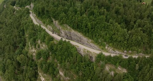 Aerial view of a winding mountain road cutting through dense green forest and rocky cliffs. Several