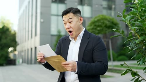 Excited asian businessman in formal suit reading letter with great news standing on the street near