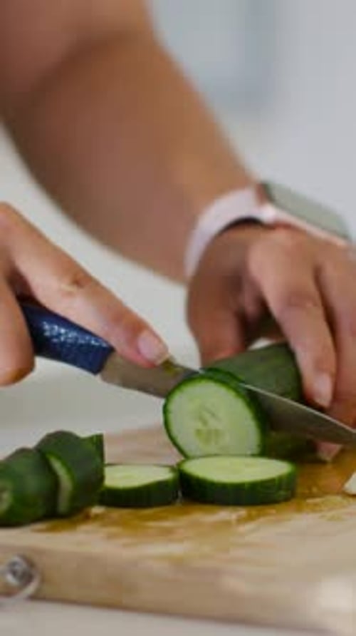 Vertical video: Hands slicing cucumber in kitchen left steadying, blue knife pink watch for salad