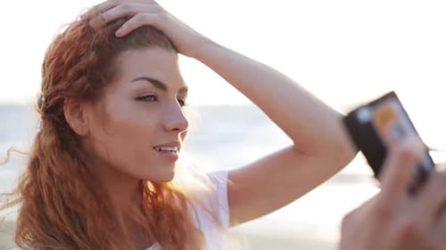 Happy young redhead woman taking a summer selfie on the beach with her smartphone