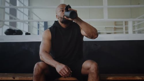 Muscular Black Athlete Drinks Water Sitting on Gym Bench