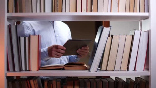 Professor Using Tablet and Consulting Book in Library