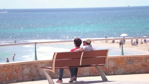 Couple of old retired seniors man and woman siting at a bench watching ocean by the beach and laughi